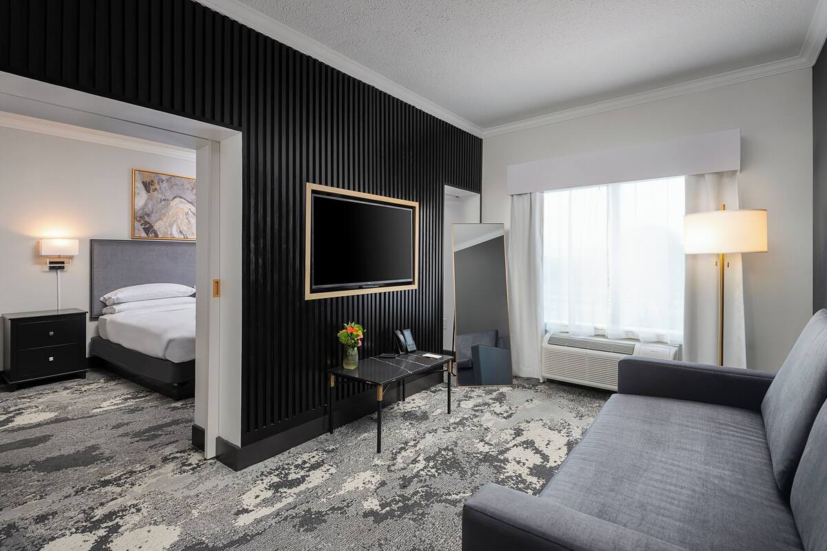 A modern hotel room with a bed, a desk with a laptop, a sofa, and a large window with curtains.