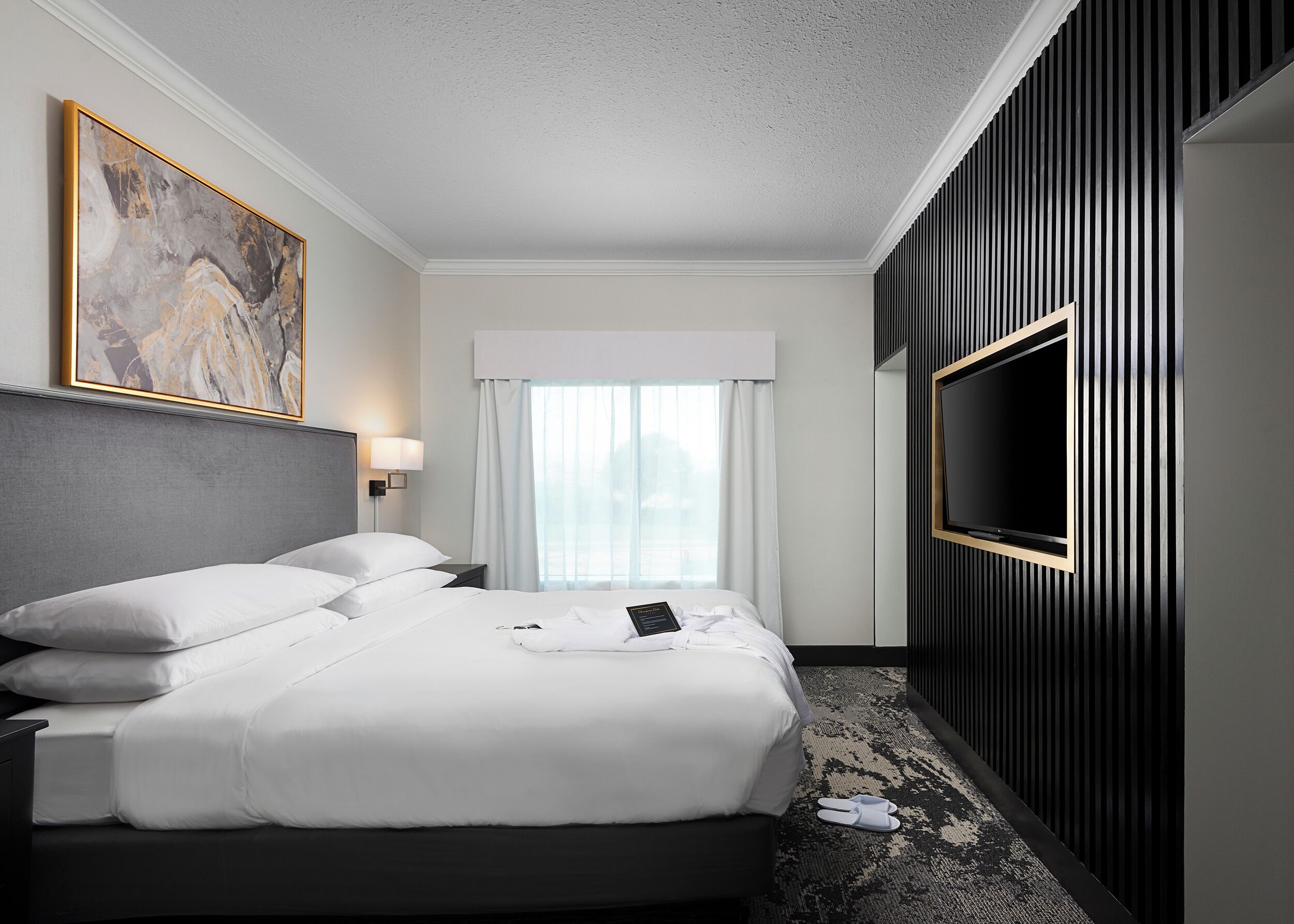 A hotel room with a large bed, a flat-screen TV mounted on the wall, a painting on the wall, and a window with curtains.