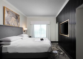 A hotel room with a large bed, a flat-screen TV mounted on the wall, a painting on the wall, and a window with curtains.