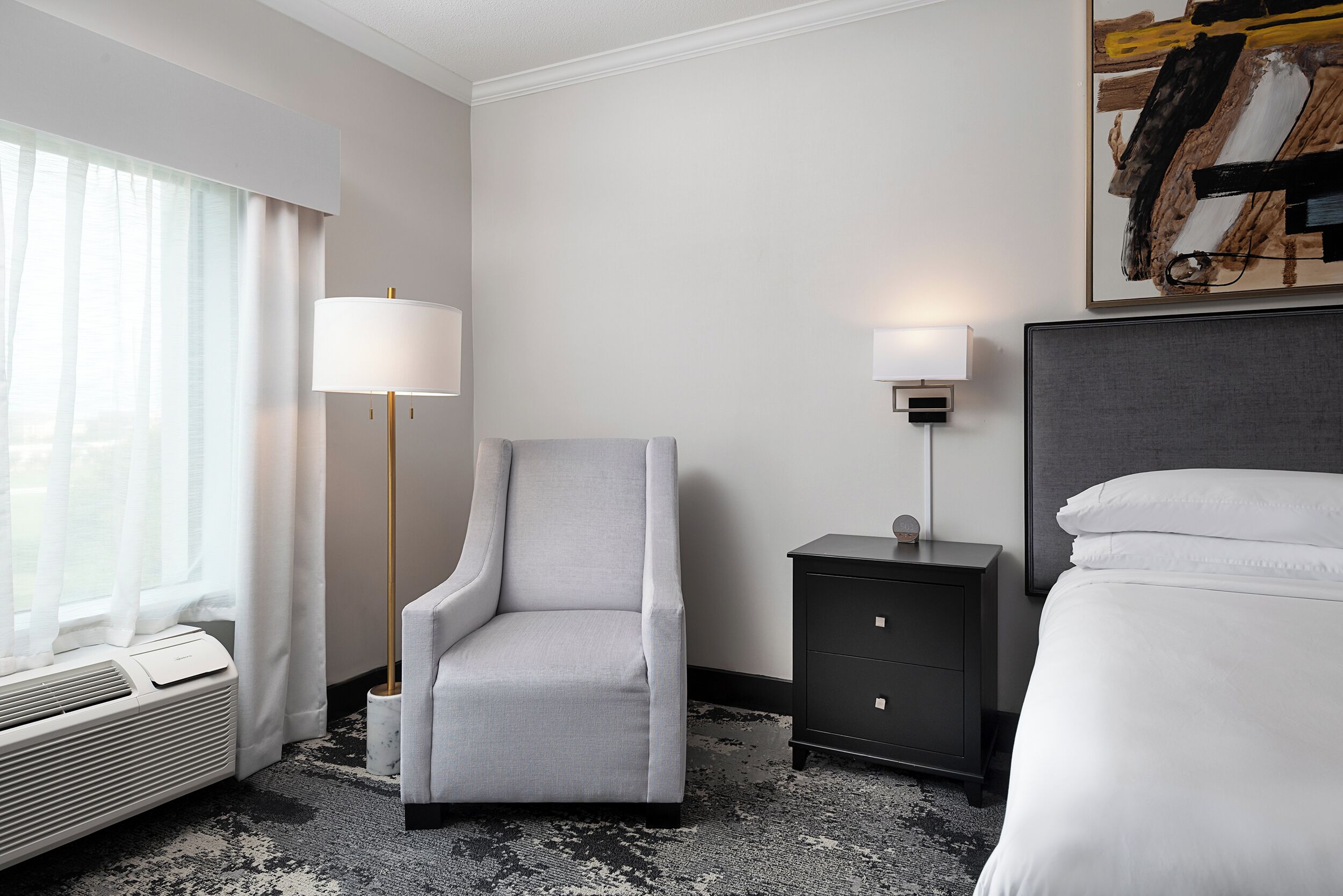 A hotel room with a bed, a chair, a nightstand, a lamp, and a painting on the wall.