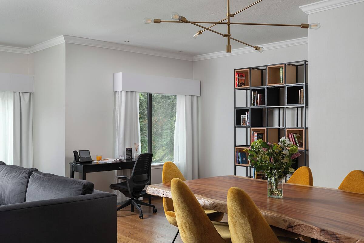 A modern dining area with a wooden table, yellow chairs, a black office chair, and a bookshelf.