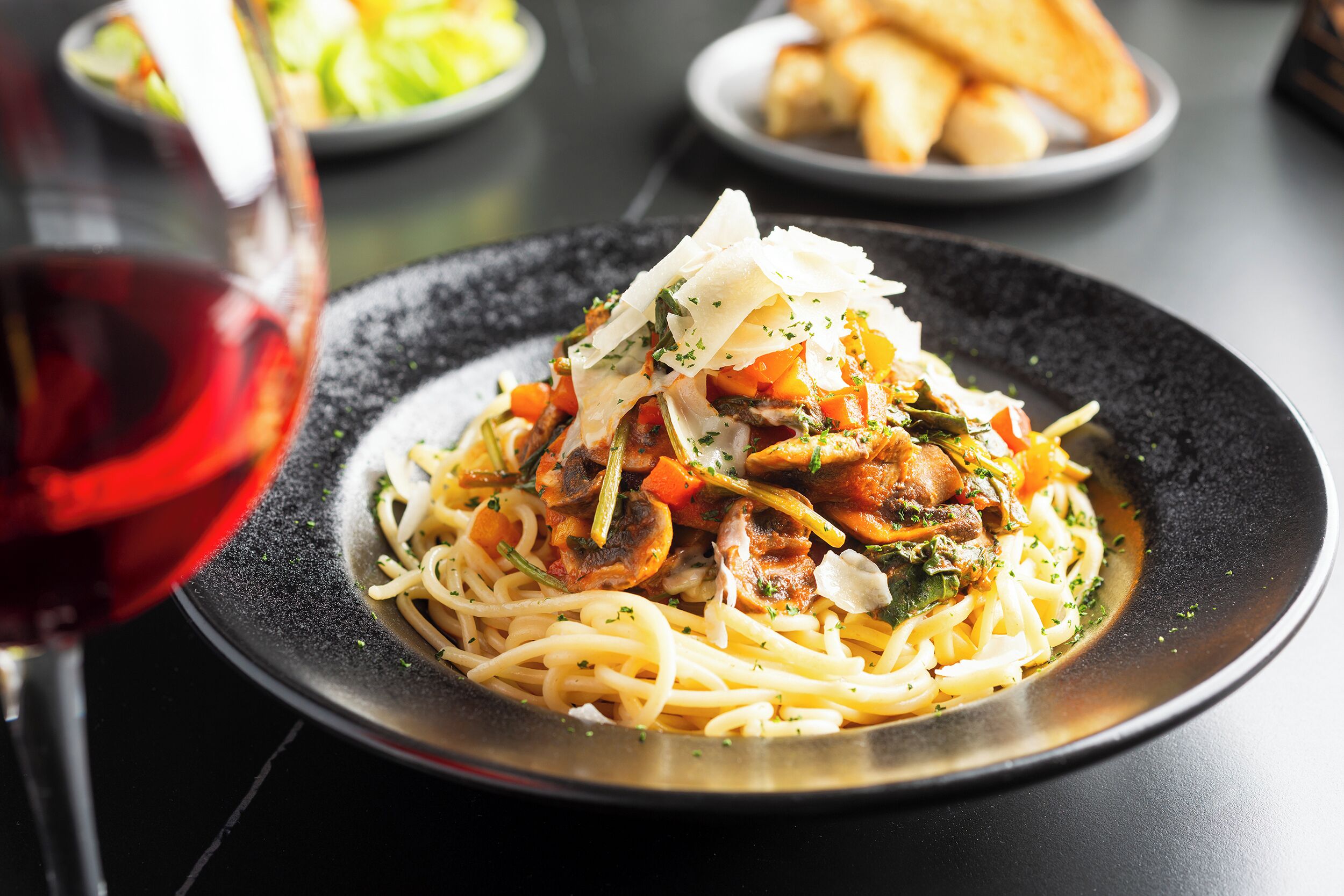 A plate of spaghetti with meat and vegetables, a glass of red wine, and bread on a table.