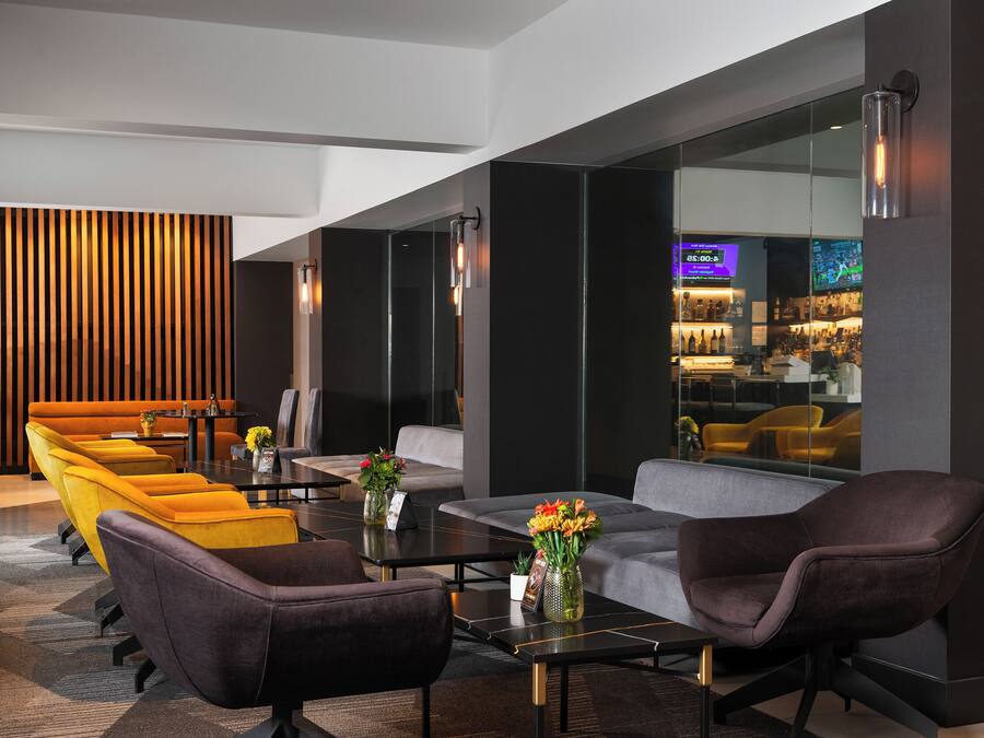 A modern lobby with a seating area featuring yellow and gray sofas, a coffee table, and a bar area with a neon sign.