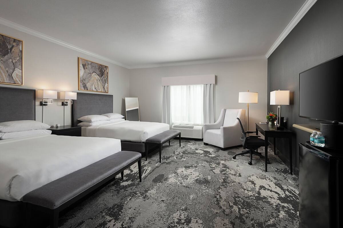 A hotel room with two beds, a desk, a chair, a TV, and artwork on the walls.