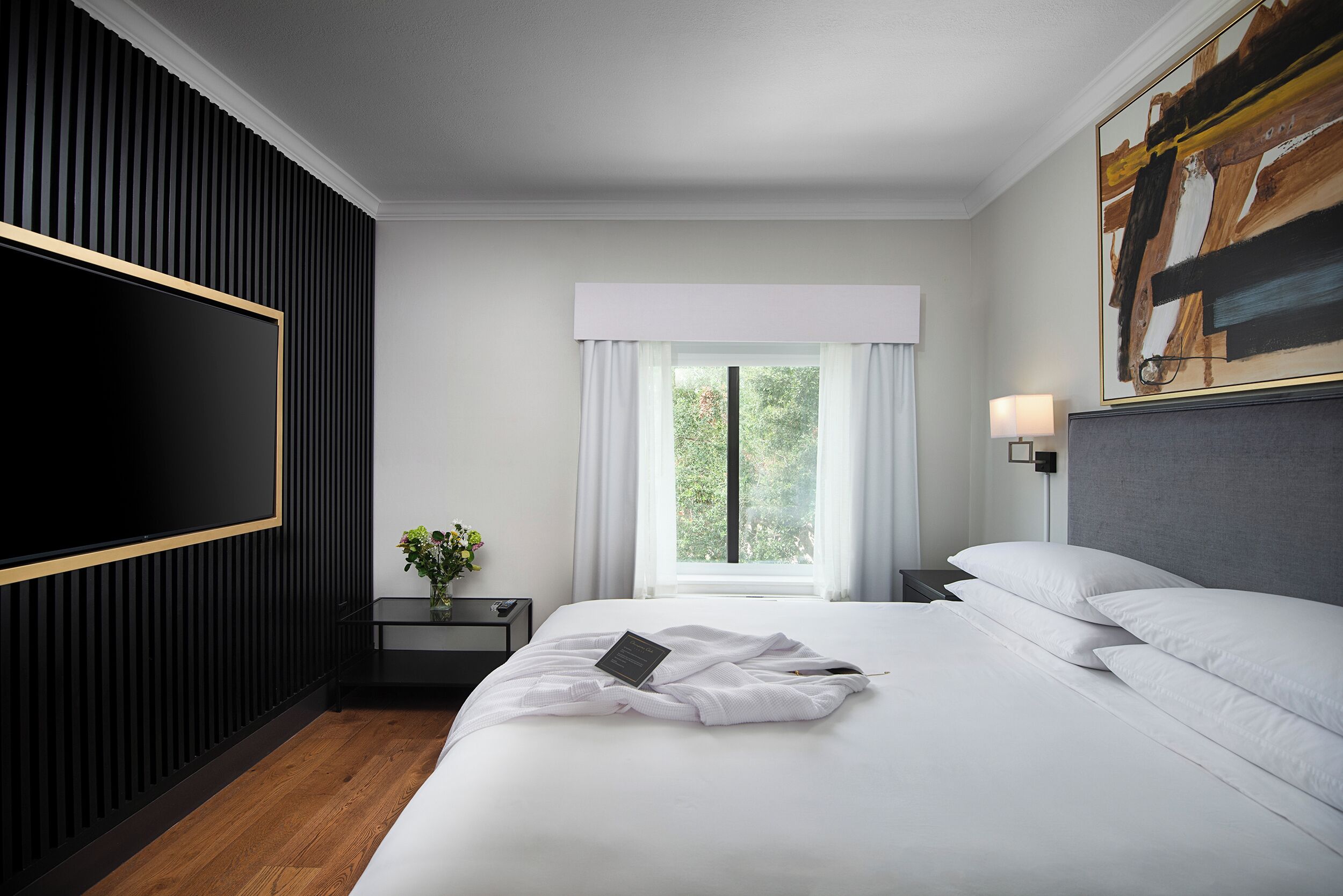 Una habitación de hotel moderna con una cama grande, un televisor de pantalla plana, una mesita de noche con lámpara y una ventana con cortinas.