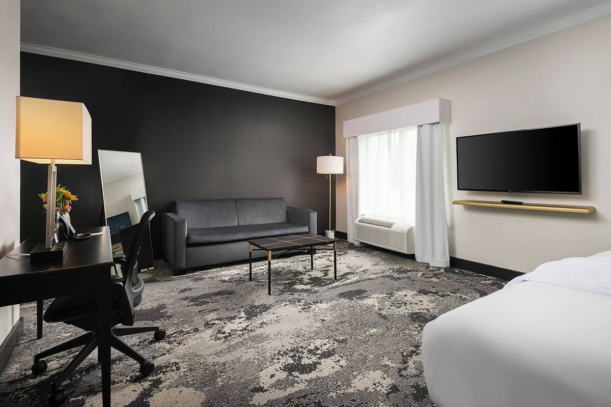 A hotel room with a grey sofa, a desk with a lamp, a flat-screen TV, and a large bed.