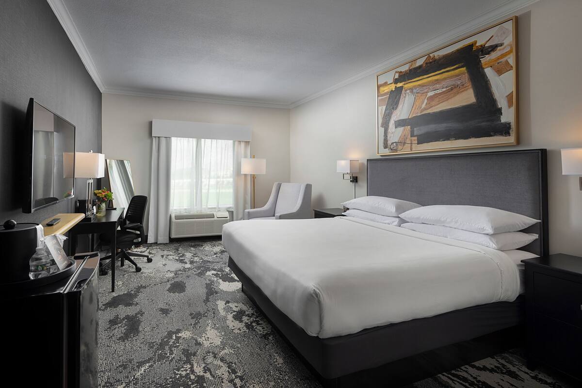 A hotel room with a large bed, a desk with a computer, a chair, and a desk lamp.