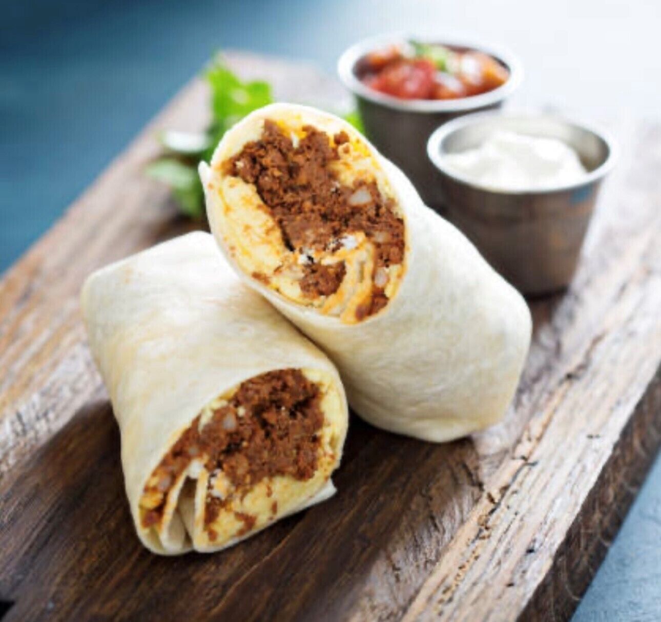 Burritos on a wooden board with a side of salsa and a small bowl of sauce.