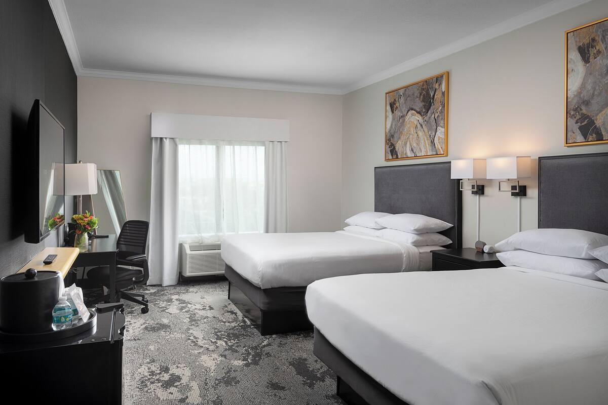 A hotel room with two beds, a desk with a lamp, a chair, a TV, and artwork on the walls.