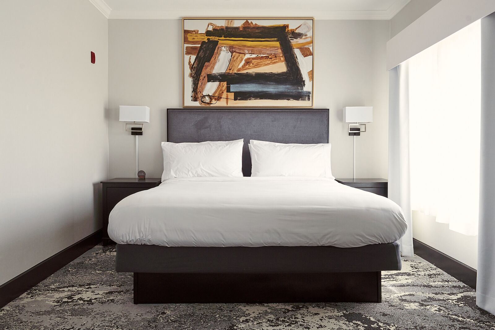Une chambre d’hôtel avec un grand lit, deux tables de chevet, une peinture abstraite moderne et une grande fenêtre avec des rideaux.