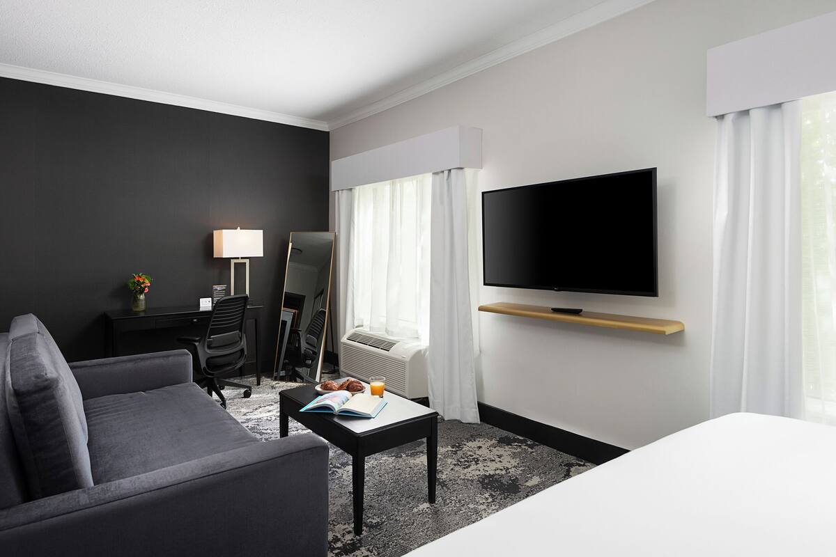 A modern hotel room with a grey sofa, a small table with a book and a glass of orange juice, a flat-screen TV mounted on the wall, and a desk with a lamp and a chair.