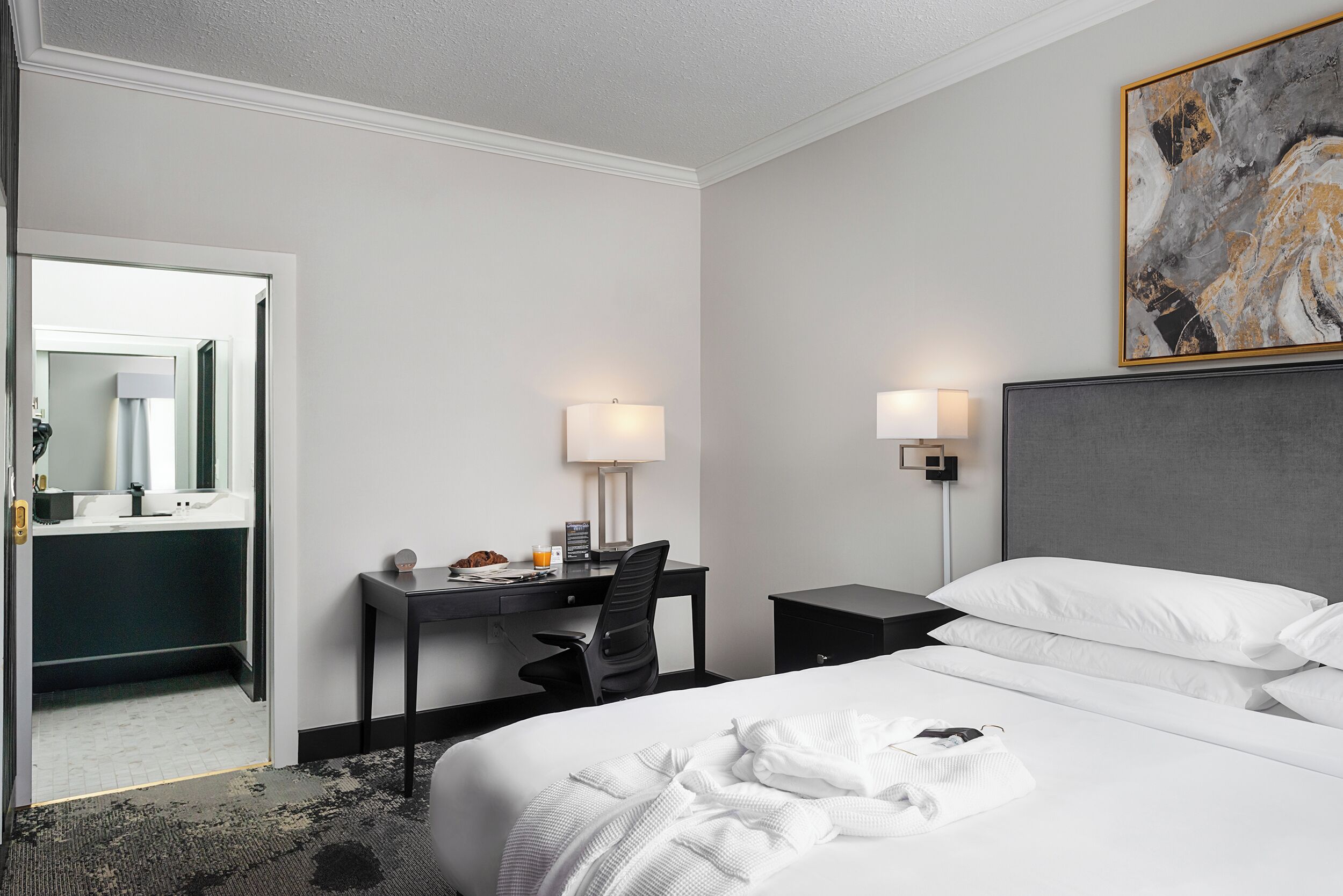 Une chambre d’hôtel comprenant un lit, un bureau, une chaise, une table de chevet et une lampe fixée au mur.