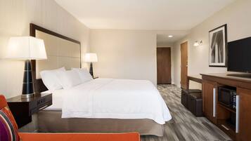 A hotel room with a large bed, bedside lamps, a television, a desk with a microwave, and a suitcase.