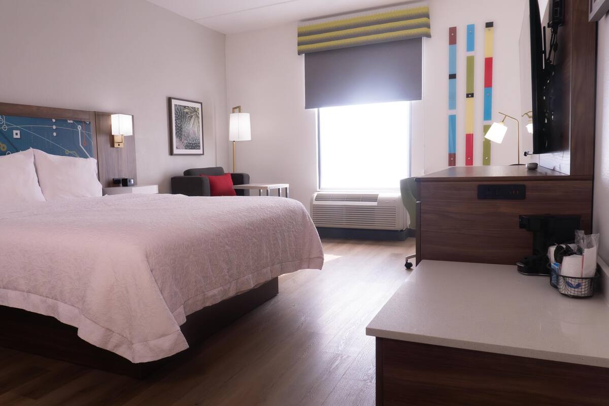 A hotel room with a bed, bedside tables, a desk, a chair, a window with a view, and an air conditioning unit.
