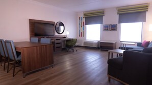 A modern living room with a wooden dining table, a green office chair, a black sofa, and a TV mounted on the wall.
