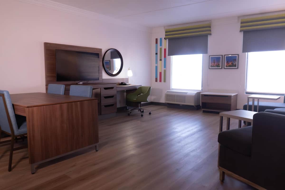 A modern living room with a wooden dining table, a green office chair, a black sofa, and a TV mounted on the wall.