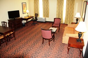 Habitación de hotel con alfombra roja estampada, una televisión, un escritorio con una silla, un sofá y dos lámparas.