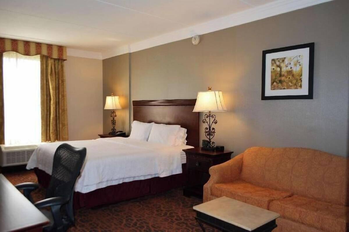 A hotel room with a bed, two bedside lamps, a framed picture on the wall, a sofa, and a desk with a chair.