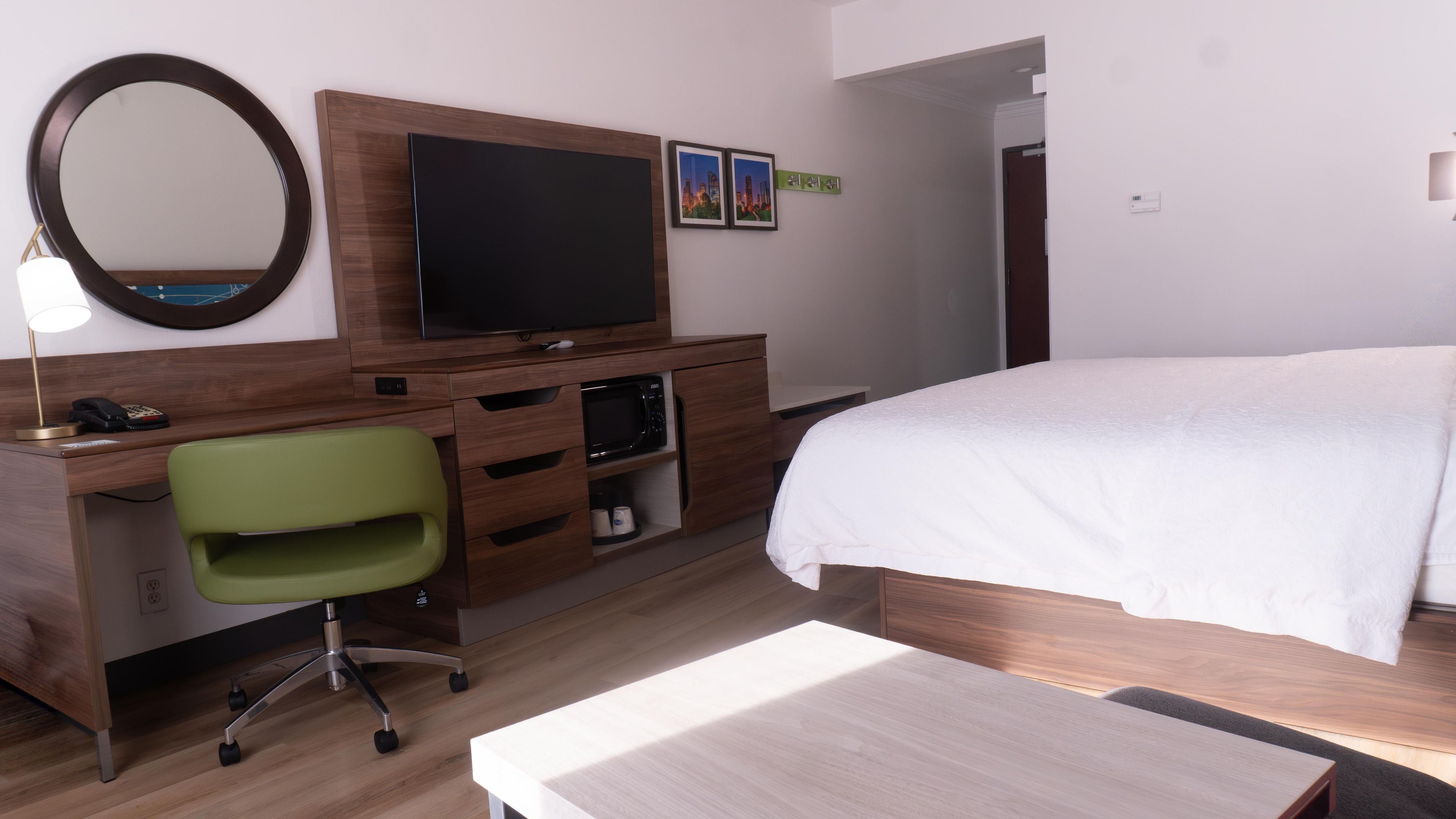 A hotel room with a bed, a desk with a chair, a television, a mirror, and a small shelf with a microwave.