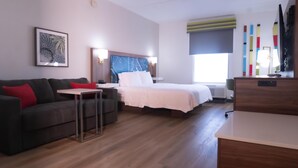 A modern hotel room with a bed, a sofa, a small table, and a desk. There is a window with blinds, a wall clock, and a framed picture.