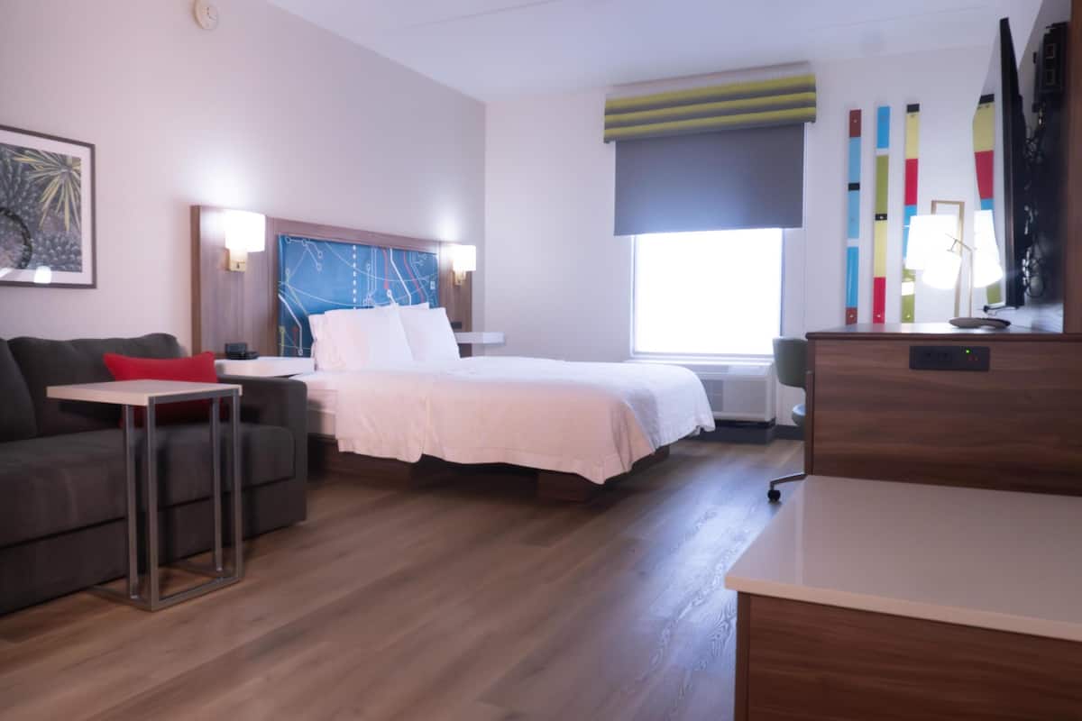 A modern hotel room with a bed, a sofa, a small table, and a desk. There is a window with blinds, a wall clock, and a framed picture.