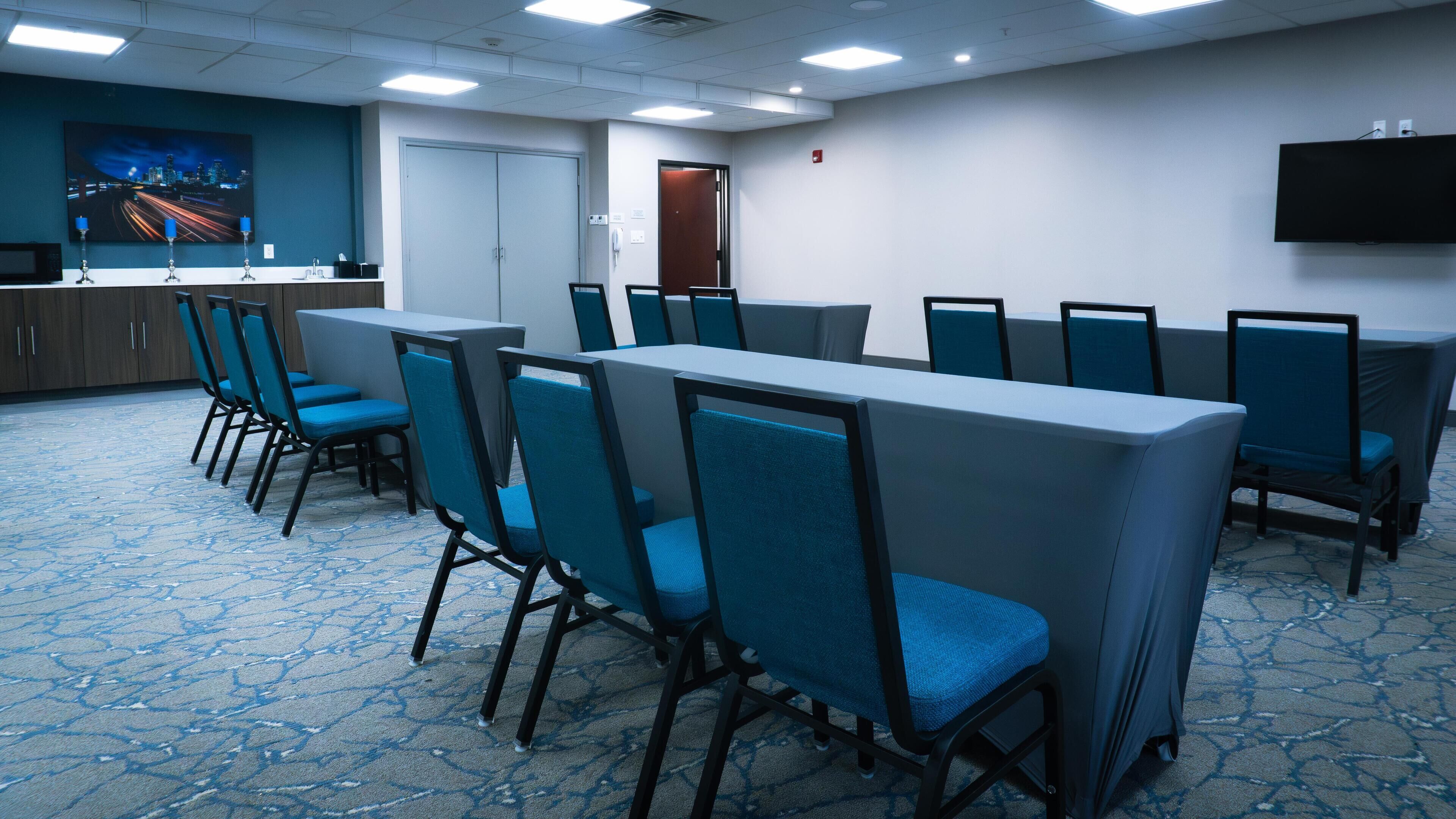 Une salle de conférence avec des rangées de chaises bleues, une table blanche et un téléviseur à écran plat accroché au mur.
