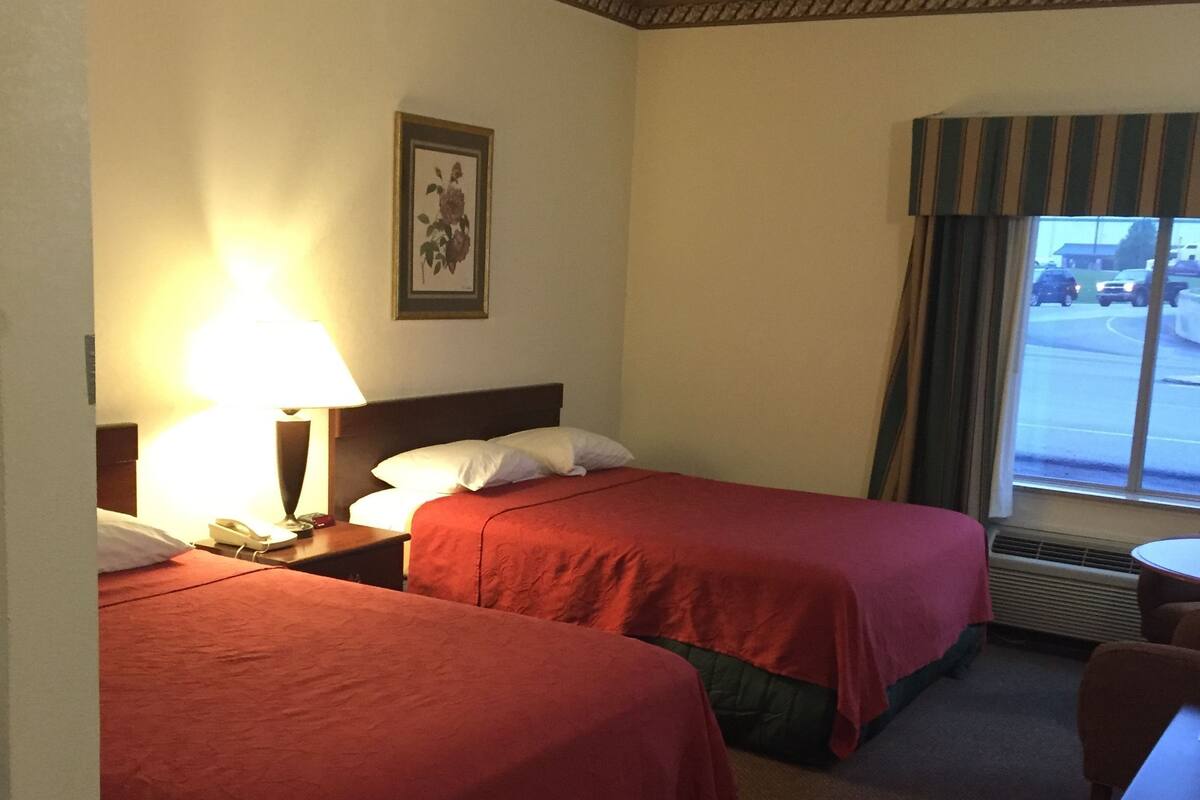A hotel room with two beds, a desk, a lamp, a framed picture on the wall, and a window with a view of the parking lot.