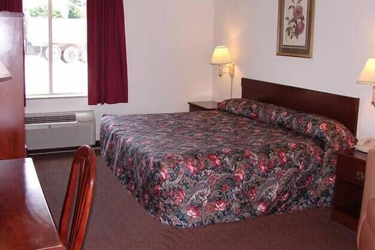 A hotel room with a bed, two bedside lamps, a window with curtains, a radiator, and a framed picture on the wall.