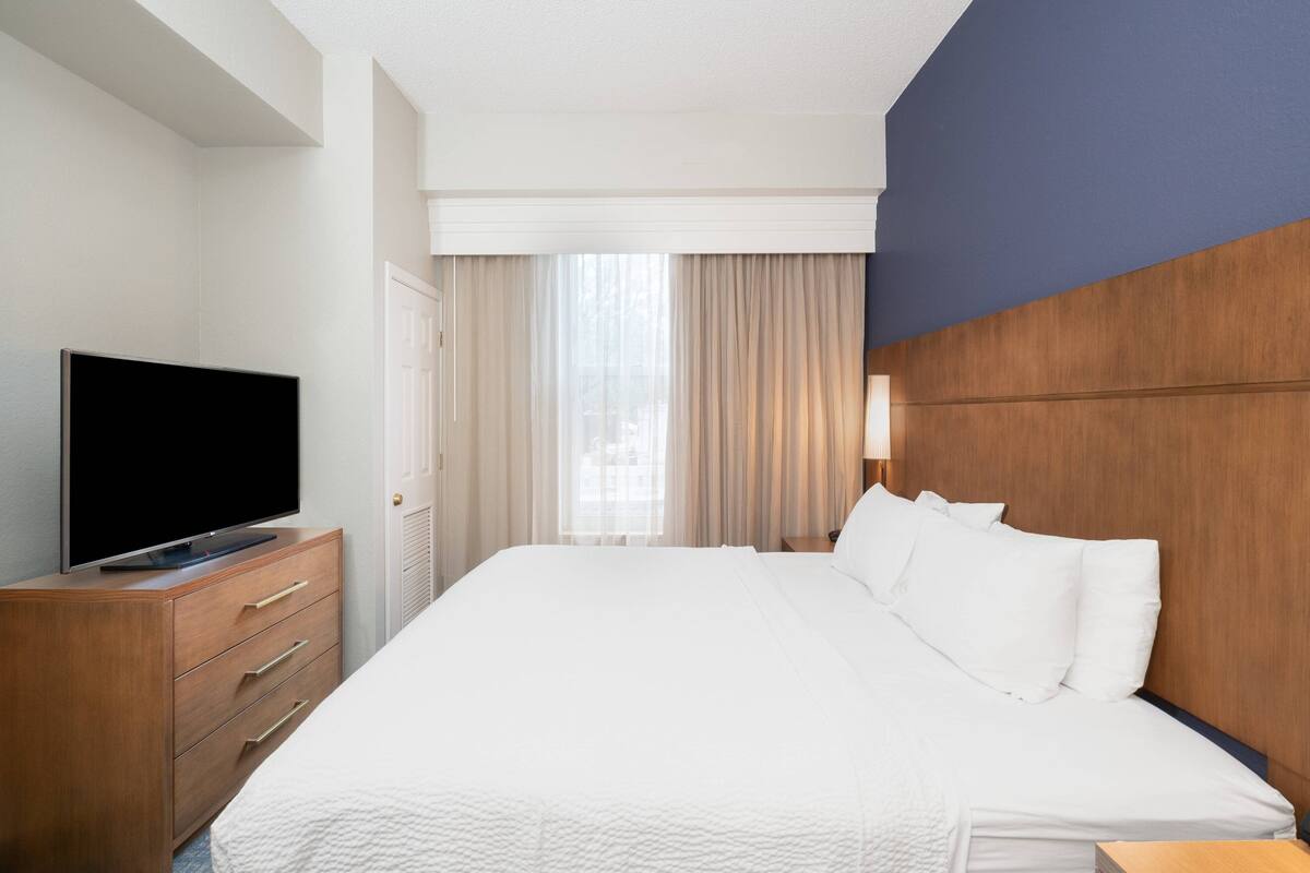 A hotel room with a large bed, a flat-screen TV on a wooden dresser, a wooden headboard, and a window with curtains.