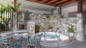 Un espace extérieur avec un jacuzzi en pierre, agrémenté d’une table et de chaises, entouré de plantes en pot et d’une clôture en métal.