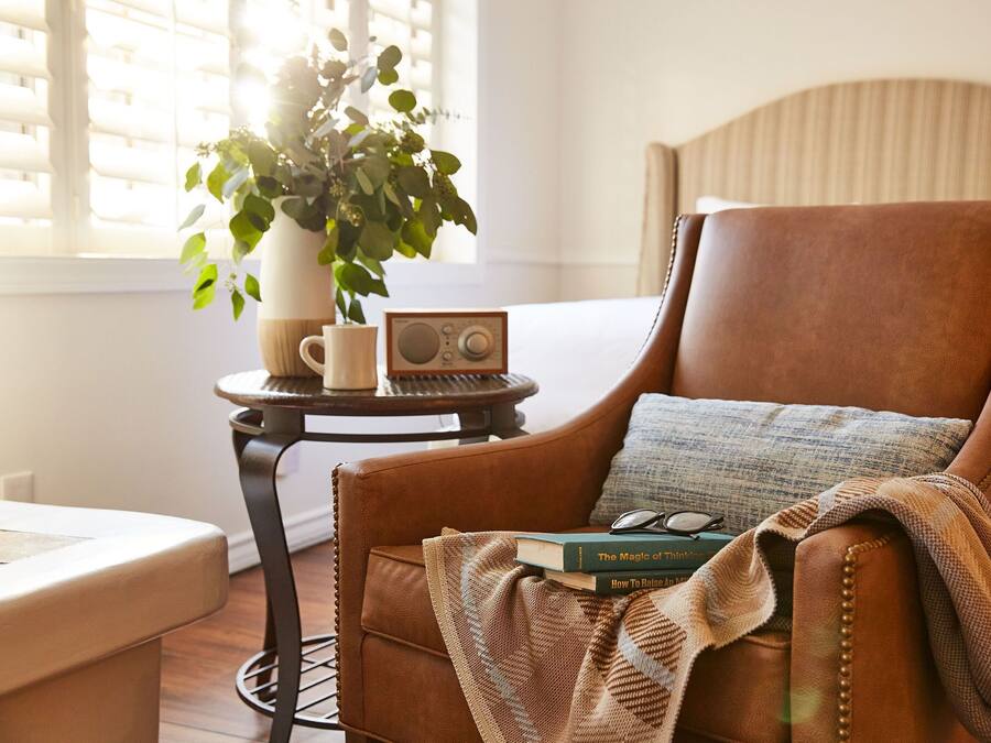 A cozy room with a leather armchair, a small round table with a radio, a potted plant, and a bookshelf with books.