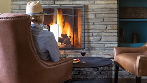 Un salon chaleureux avec une cheminée en pierre, des fauteuils en cuir et une petite table ronde sur laquelle repose un verre de vin.