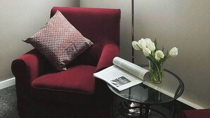 A red armchair with a patterned pillow, a floor lamp, a glass table with a magazine and a vase of white flowers. - Teledom Hotel & Conference Center (Kosice)