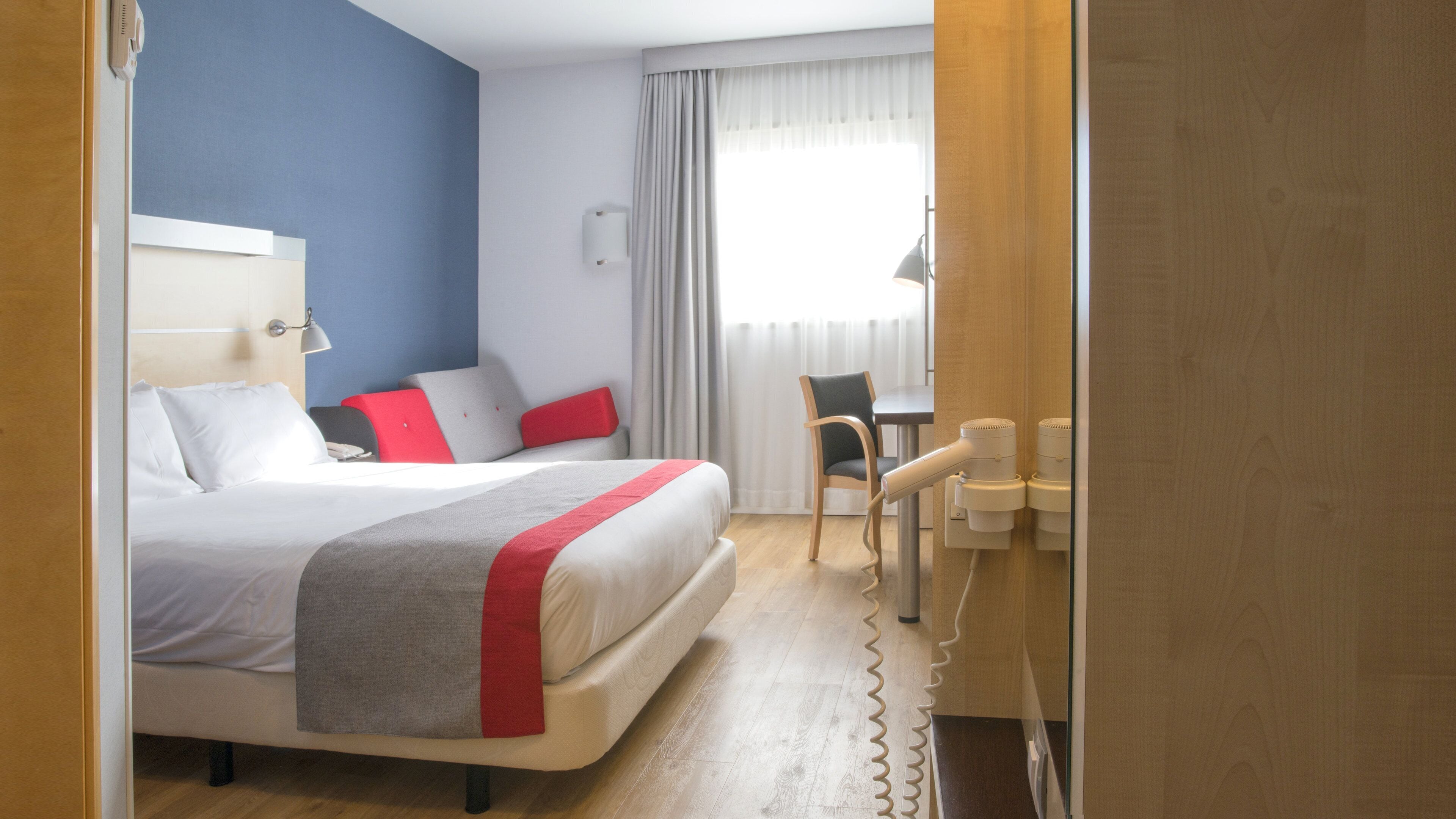 Quarto de hotel com cama, escrivaninha, cadeira e janela com cortinas.