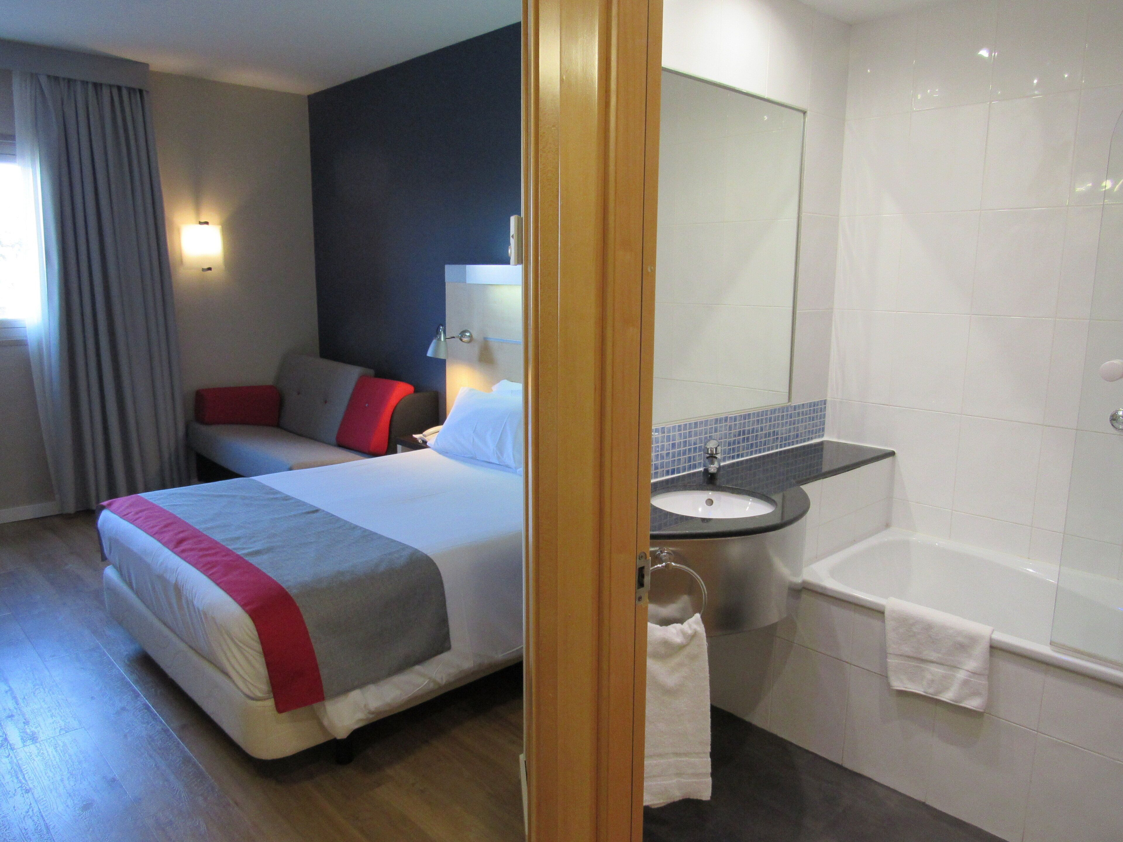 A hotel room with a bed, a bedside table, a lamp, a bathroom with a sink, and a bathtub.