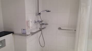 Une salle de bain avec des toilettes blanches, une douche équipée d’un pommeau de douche amovible et d’une barre d’appui.