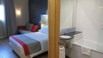 Habitación de hotel con cama, mesita de noche, lámpara, baño con lavamanos y bañera.