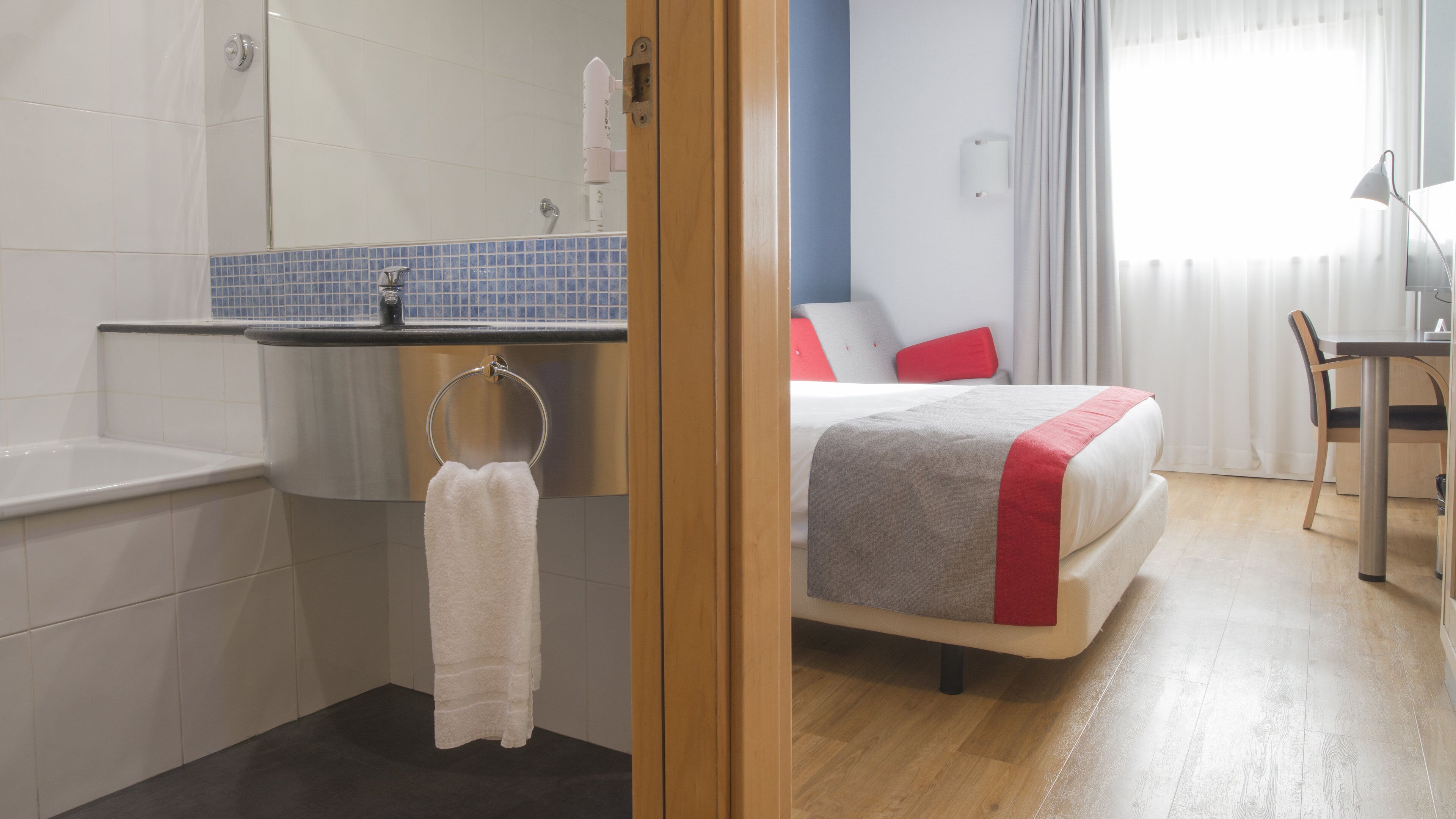 Een hotelkamer met badkamer, een bed met rode accenten en een bureau met stoel.