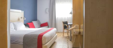 A hotel room with a bed, a desk with a chair, a lamp, and a window with curtains.