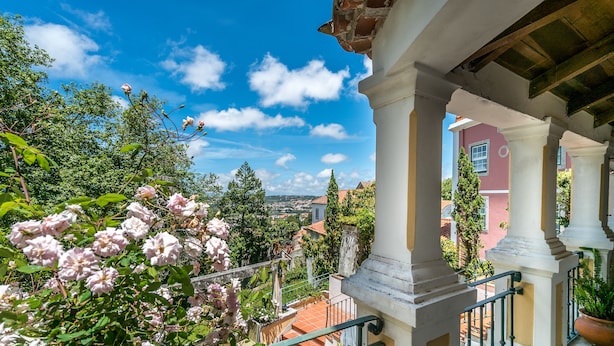 A balcony with white columns, a view of a town, and blooming pink flowers.