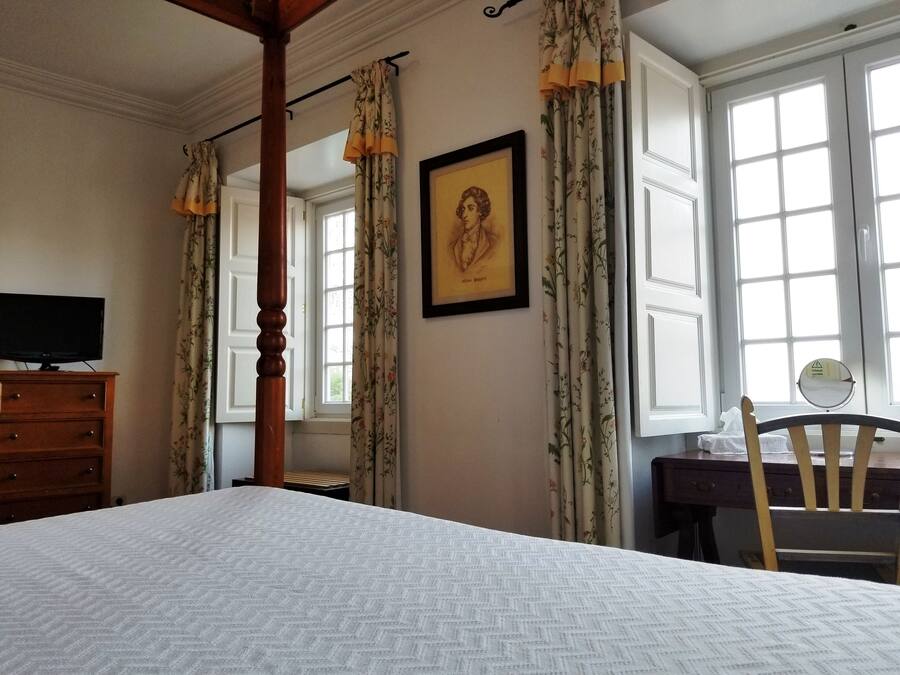 Una habitación de hotel con una cama grande, un aparador de madera, un escritorio con una silla, un cuadro enmarcado en la pared y varias ventanas con cortinas.