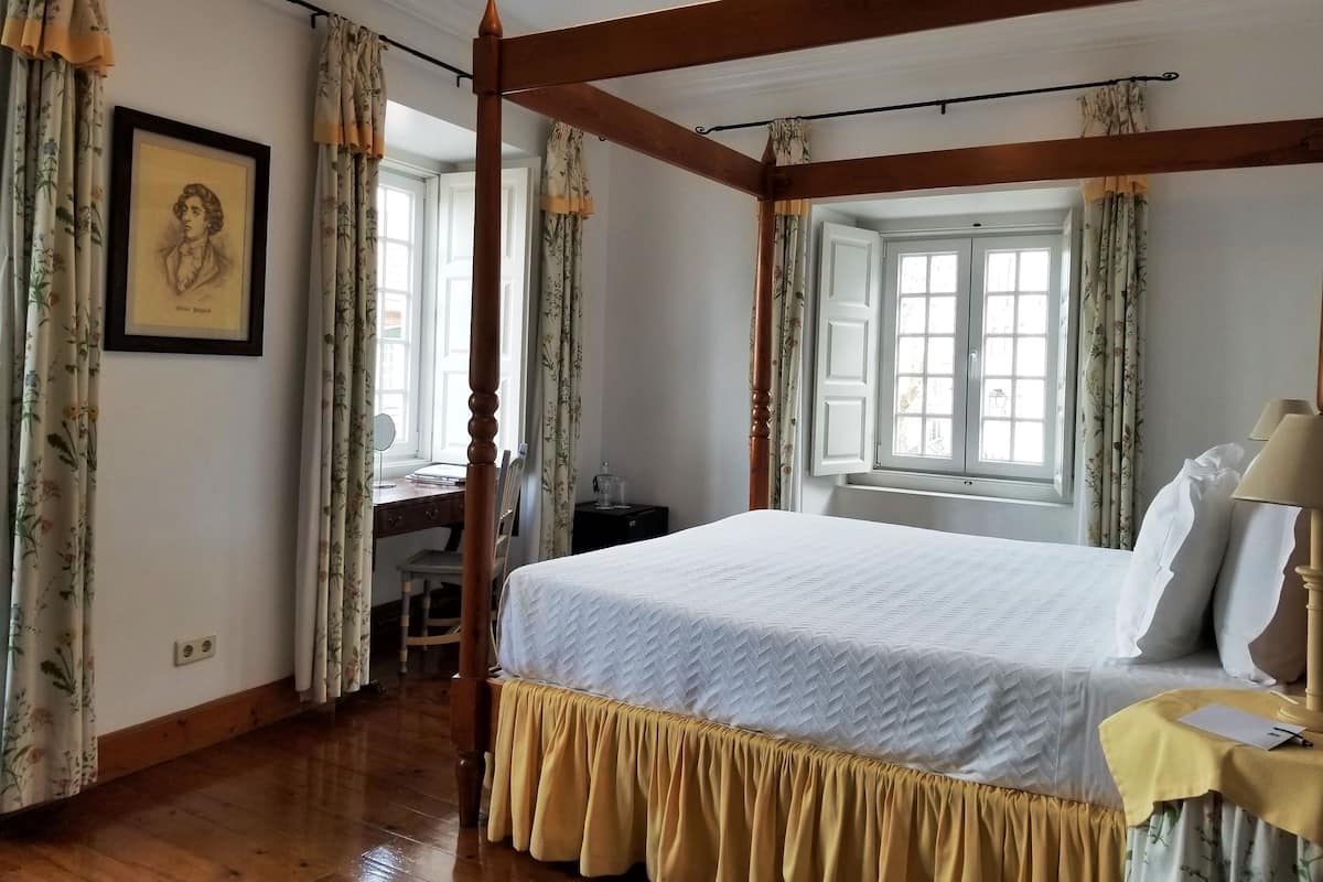 Une chambre à coucher avec un lit à baldaquin, des rideaux à motifs floraux, un bureau avec une chaise et une fenêtre avec des volets.