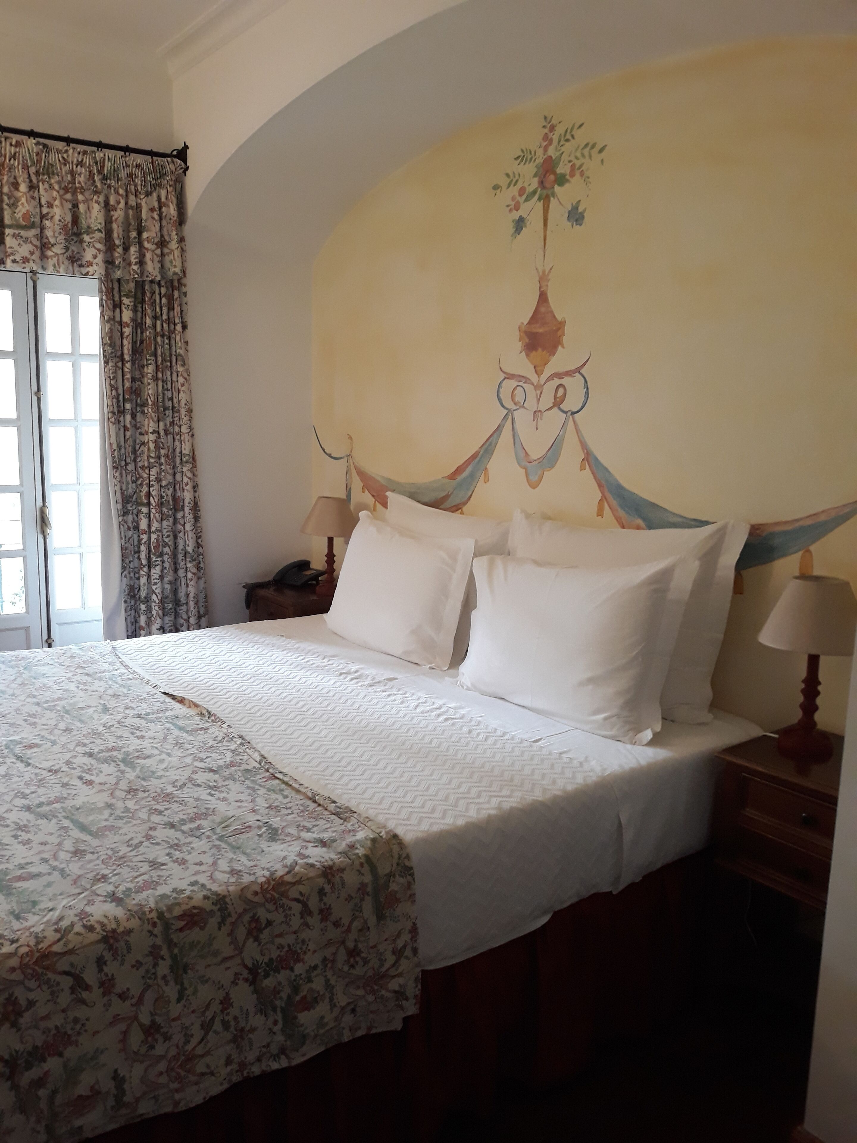 A hotel room with a large bed, a nightstand, a lamp, and a floral curtain.