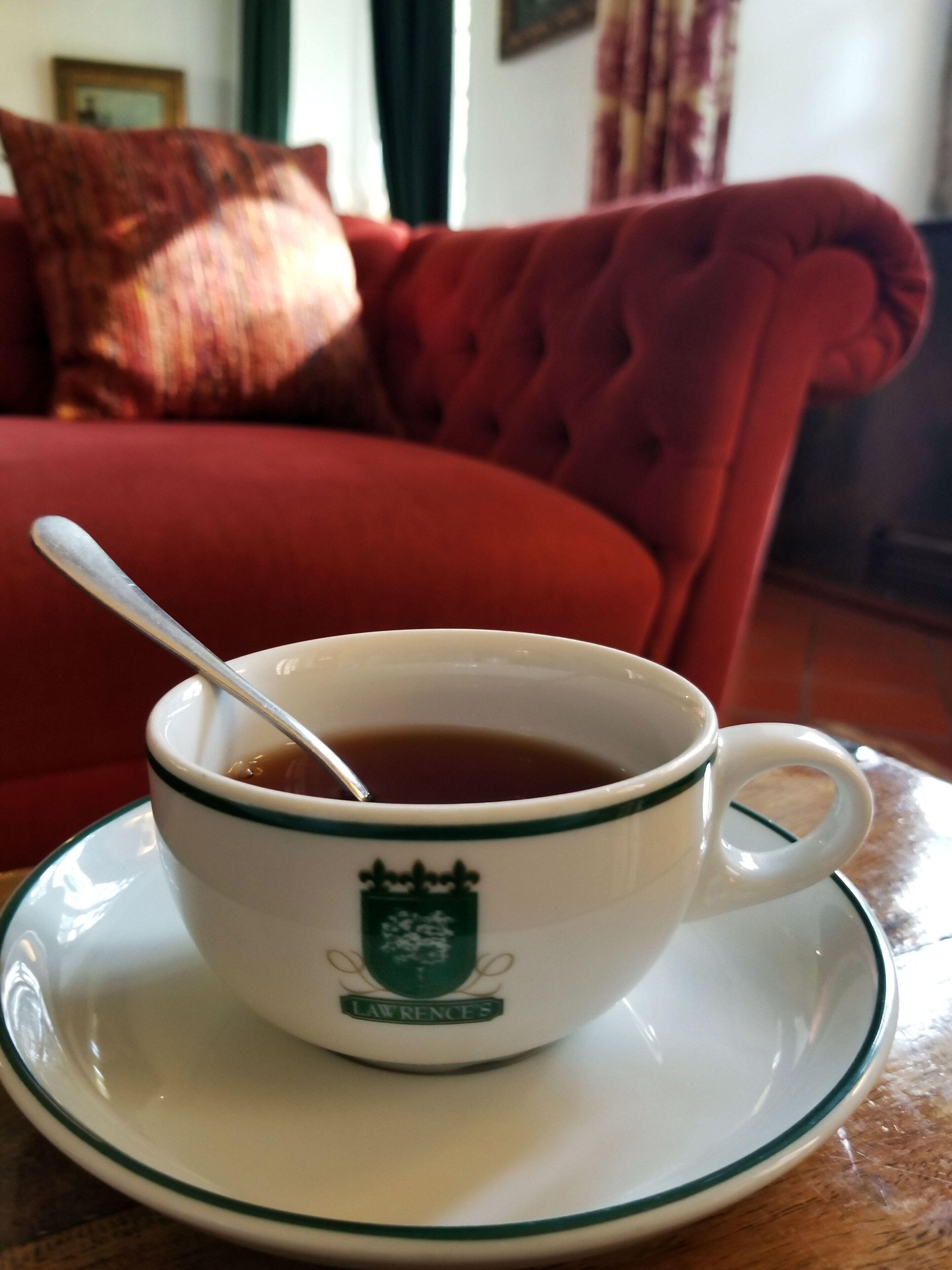 Una taza de té sobre un platito con una cuchara, sobre una mesa en una habitación con un sofá rojo y un cojín al fondo.