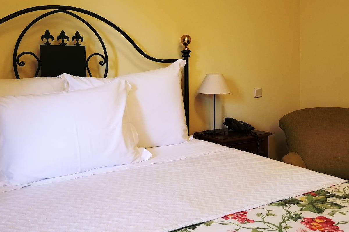 Un lit bien fait, avec du linge de lit blanc, une couvre-lit à motifs floraux, une tête de lit en métal noir ornée d’un motif décoratif, une lampe de chevet et un téléphone posés sur une table de chevet en bois.