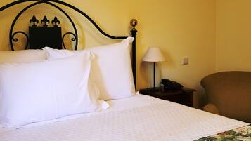 A neatly made bed with white linens, a floral-patterned bedspread, a black metal headboard with a decorative crest, a bedside lamp, and a telephone on a wooden nightstand.