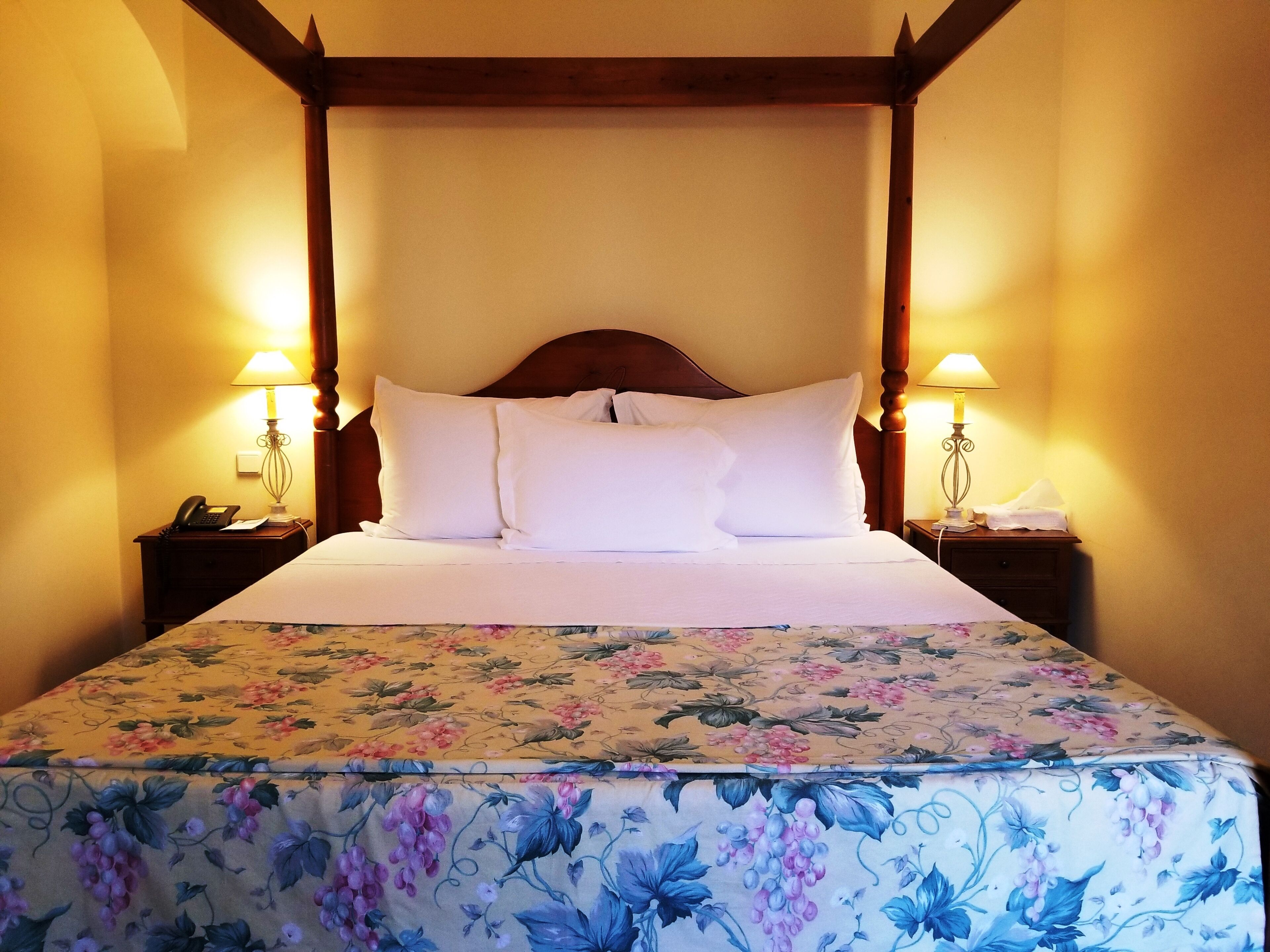 A four-poster bed with white linens and a floral bedspread. There are two bedside tables, each with a lamp and a telephone.