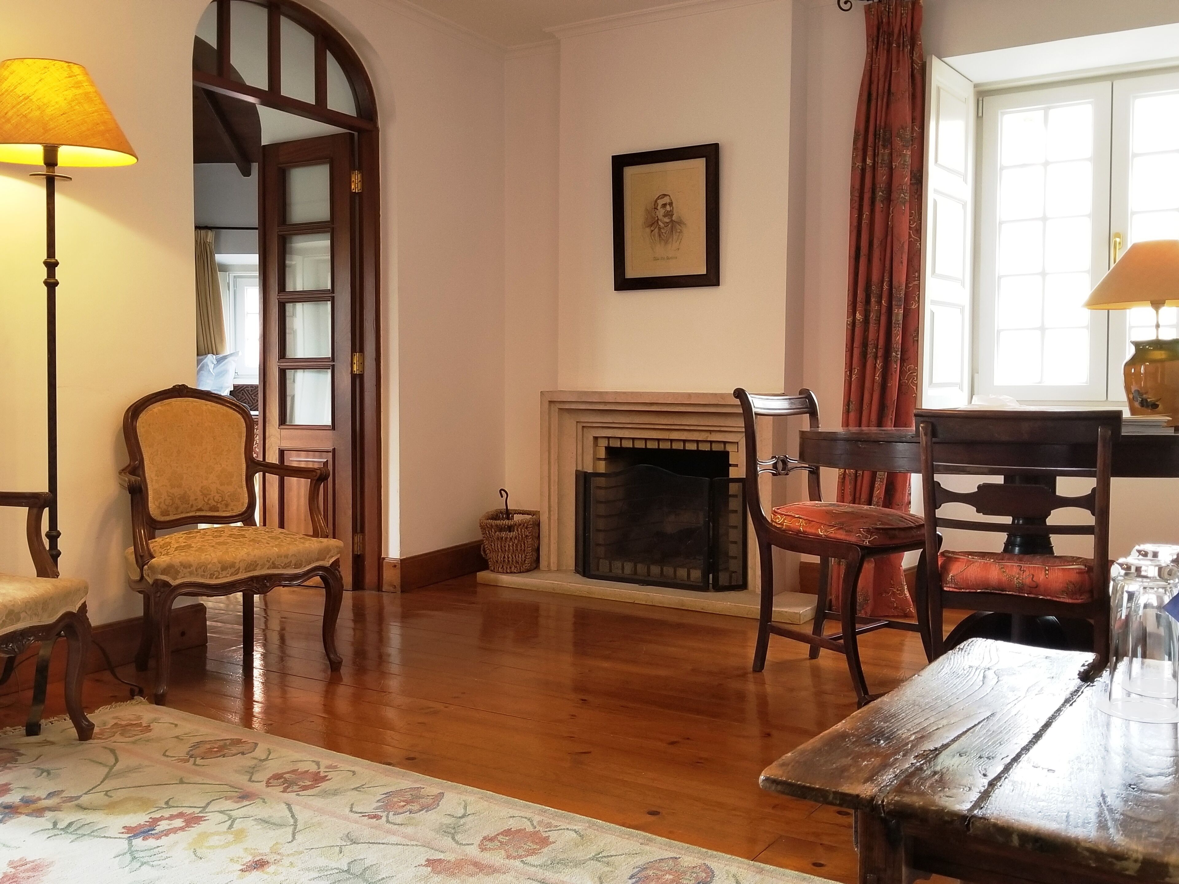 A room with a wooden floor, a fireplace, a framed portrait on the wall, and two chairs.