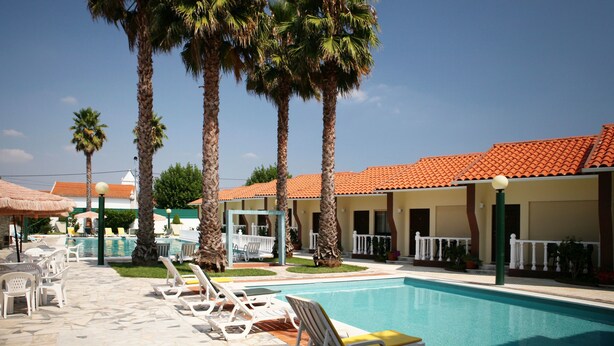 A pool area with lounge chairs, palm trees, and a building with a red-tiled roof. - Quinta dos Três Pinheiros (Mealhada)