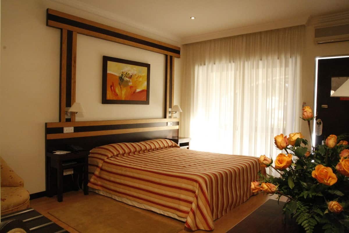 A hotel room with a bed, a nightstand, a lamp, a painting on the wall, a window with curtains, and a vase with roses.